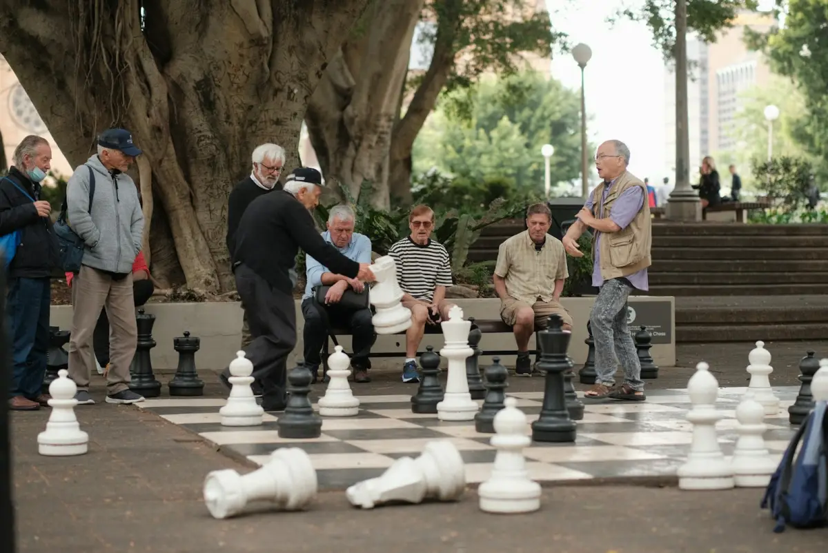 Spieler rund um ein Riesenschachbrett unter den Bäumen eines Stadtparks.